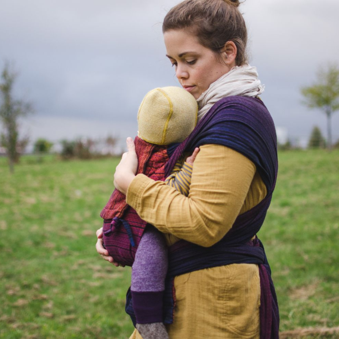 Baby in Trage, getragen von der Mama. WrapMySol Twilight. Im Hintergrund grüne Büsche